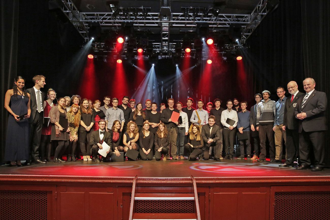 Grosse Gruppe von Menschen steht auf einer Bühne unter roten Scheinwerfern für ein gemeinsames Gruppenfoto.