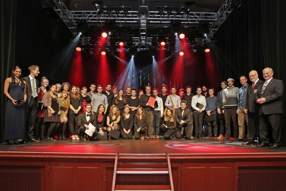 Grosse Gruppe von Menschen steht auf einer Bühne unter roten Scheinwerfern für ein gemeinsames Gruppenfoto.