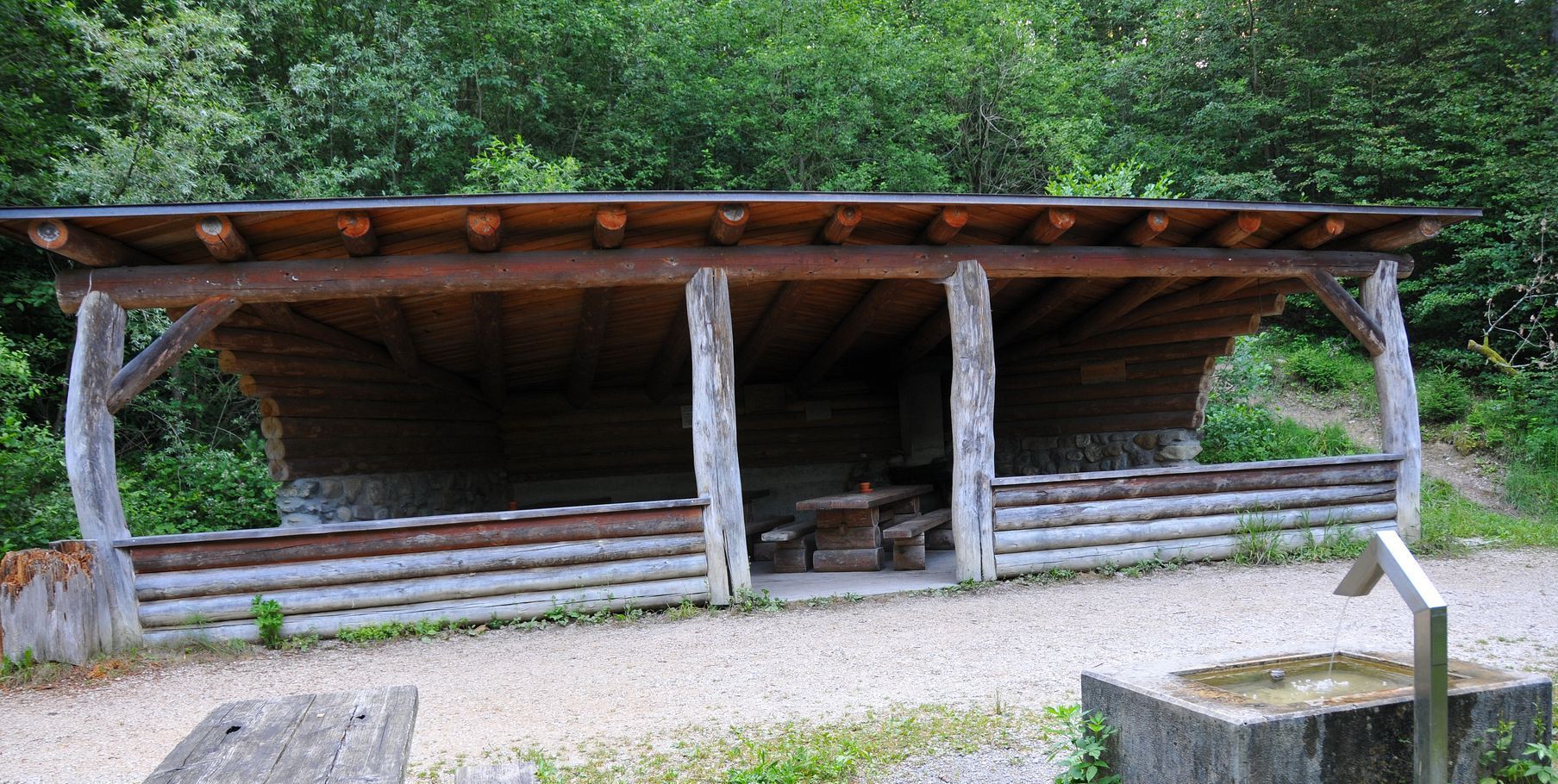 Aussenansicht der Waldhütte Spilwald mit Brunnen, Tisch und Bänken im Freien