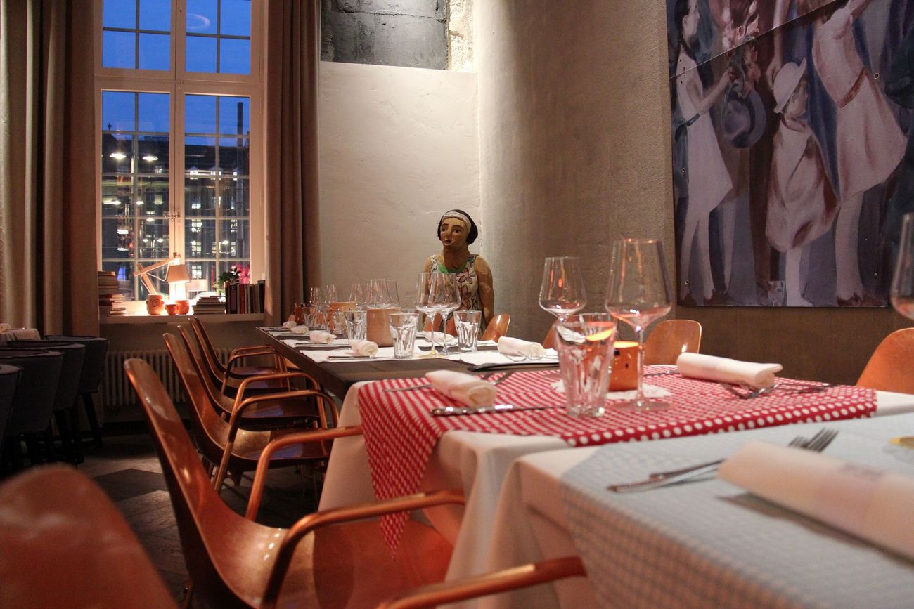 Gemütlich gedeckter Restauranttisch in einem warm beleuchteten Raum, neben einem grossen Fenster mit Abendblick auf den Bahnhofplatz.