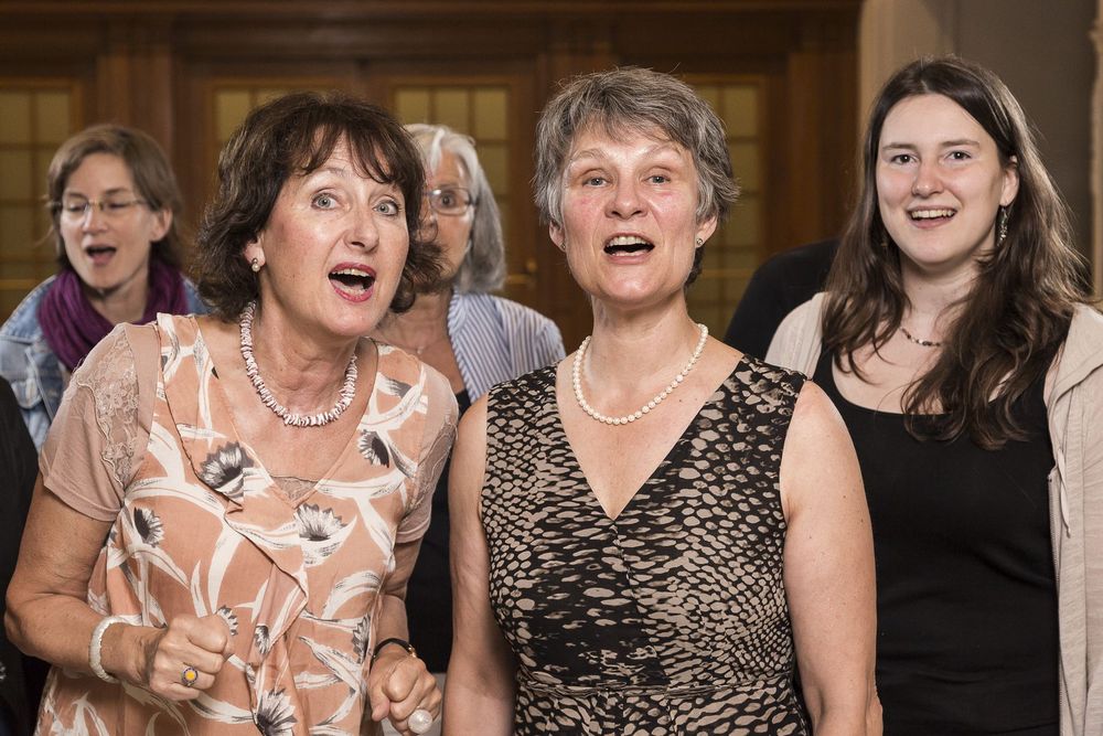 Mehrere Personen stehen dicht beisammen in einem Innenraum; zwei Frauen im Vordergrund tragen elegante Kleidung und Schmuck.