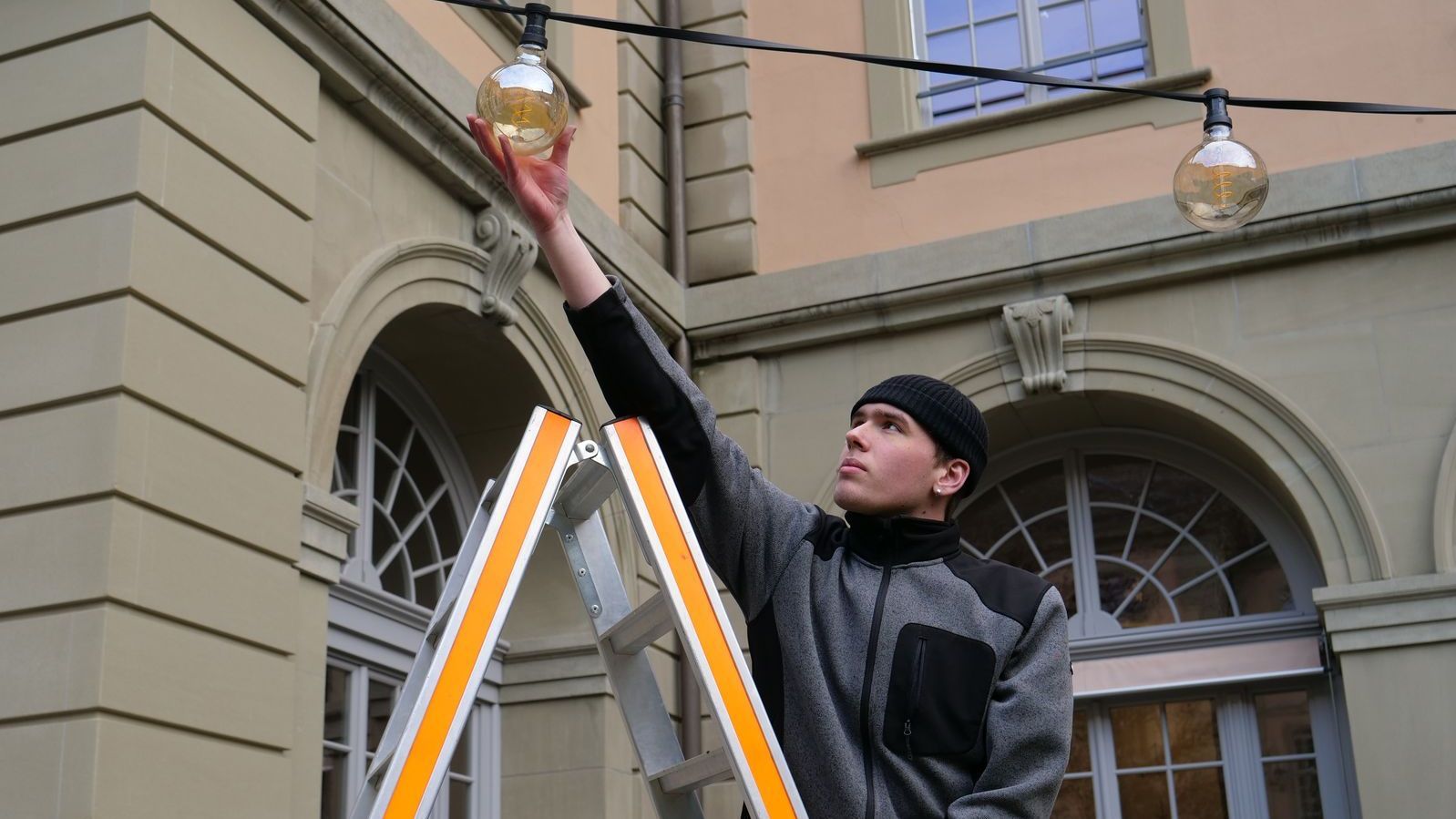 Lernender vom Hausdienst steht auf einer Leiter und hängt Glühbirne auf