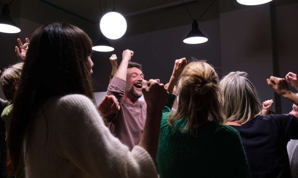 Eine Gruppe von Menschen steht eng beisammen und hebt lachend die Arme in die Höhe, als Teil einer gemeinsamen Übung. Die Szene ist warm beleuchtet und vermittelt eine lebendige, energievolle Atmosphäre.