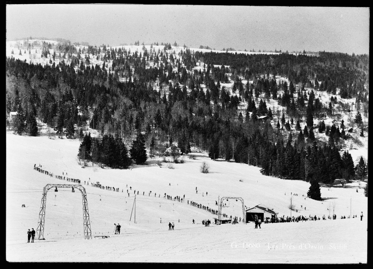 Alte Schwarz‑Weiss‑Fotografie eines verschneiten Skihangs mit vielen Menschen auf der Piste und im Waldhang dahinter.