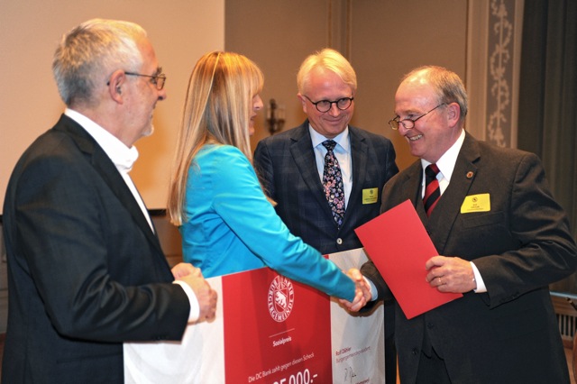 Personen bei einer feierlichen Übergabe; ein grosser Spendenscheck und ein roter Umschlag werden überreicht.